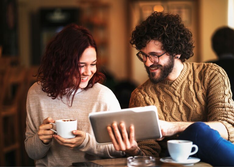 Zwei Personen sitzen an einem Tisch mit Café und schauen interessiert sowie lachend auf ein Tablet