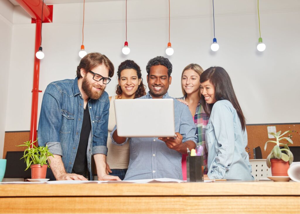 Fünf junge Menschen schauen interessiert und freundlich in einen Laptop.