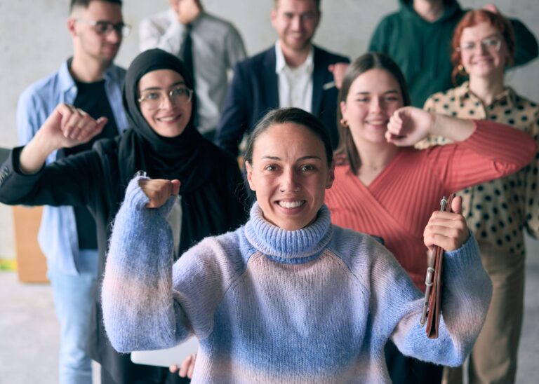 Es sind acht Menschen zu sehen, die freudig in die Kamera schauen. Die Tiefenschärfe nimmt mit Entfernung stark ab und so sind Menschen im Hintergrund unschärfer.. Manche Menschen drücken die Freude mit in die Luft gestreckten Armen aus.