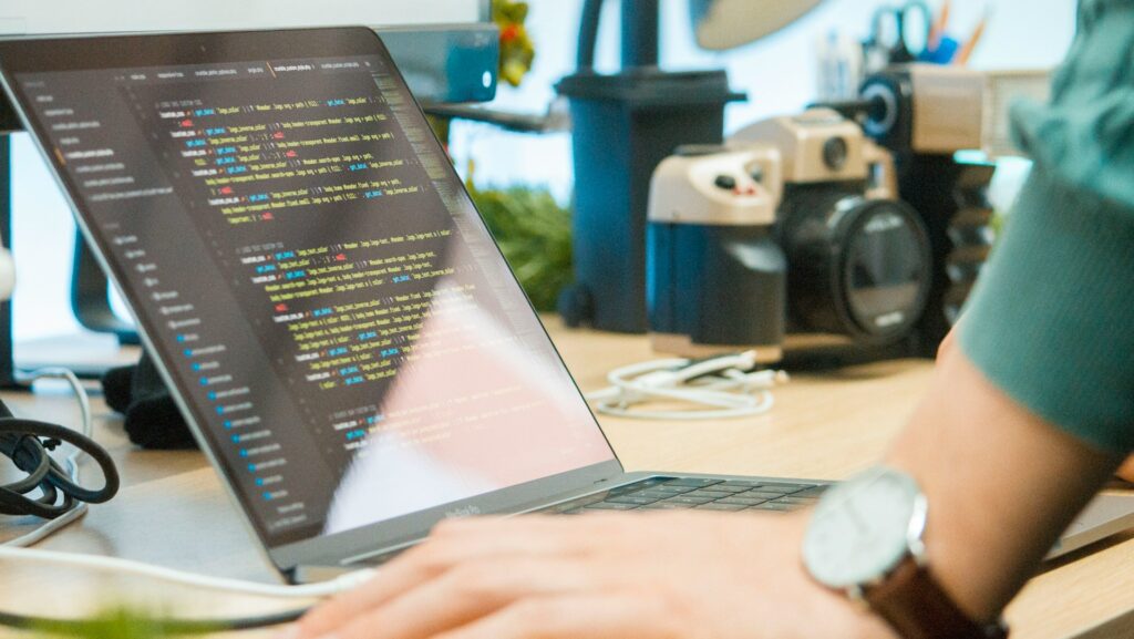 Laptop mit Code auf Tisch, im Vordergrund ein Arm mit Uhr.